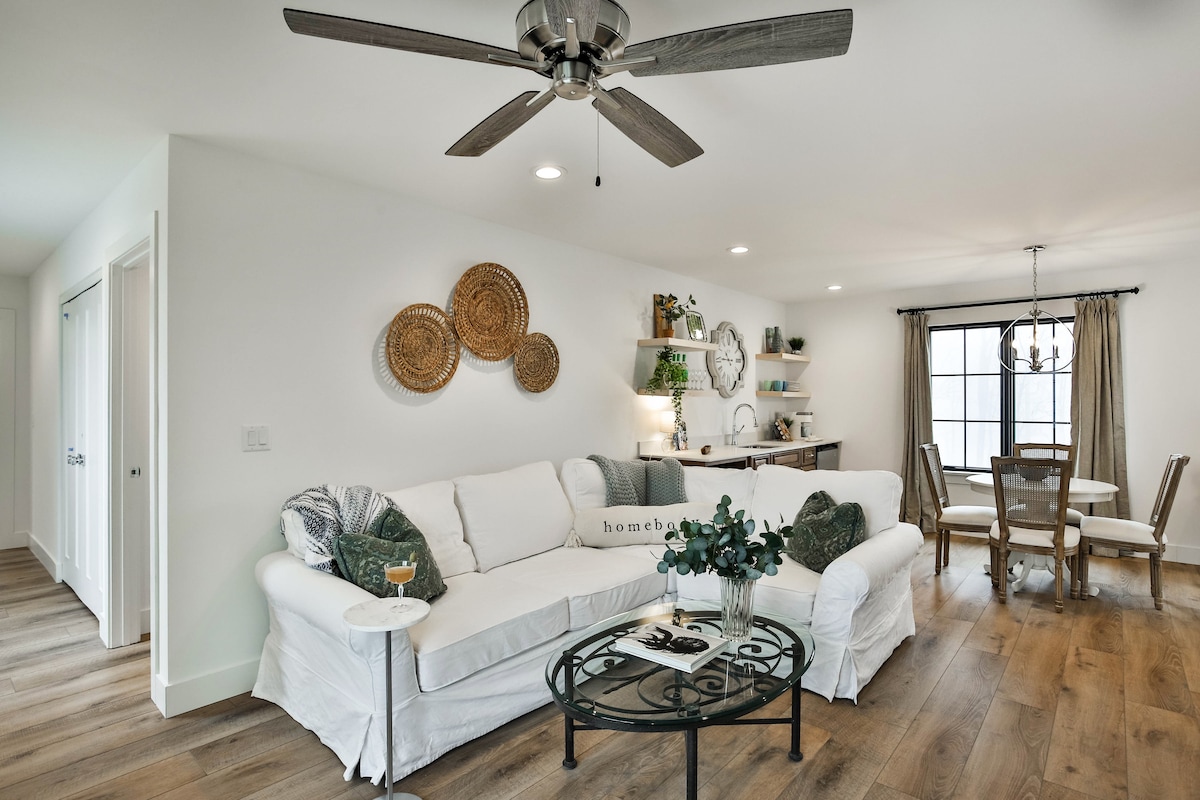 The inviting living area features a white sectional sofa adorned with green and patterned cushions. A glass coffee table sits center stage, and a ceiling fan circulates air. A dining space with a round table is visible in the background, complemented by natural light from a window.