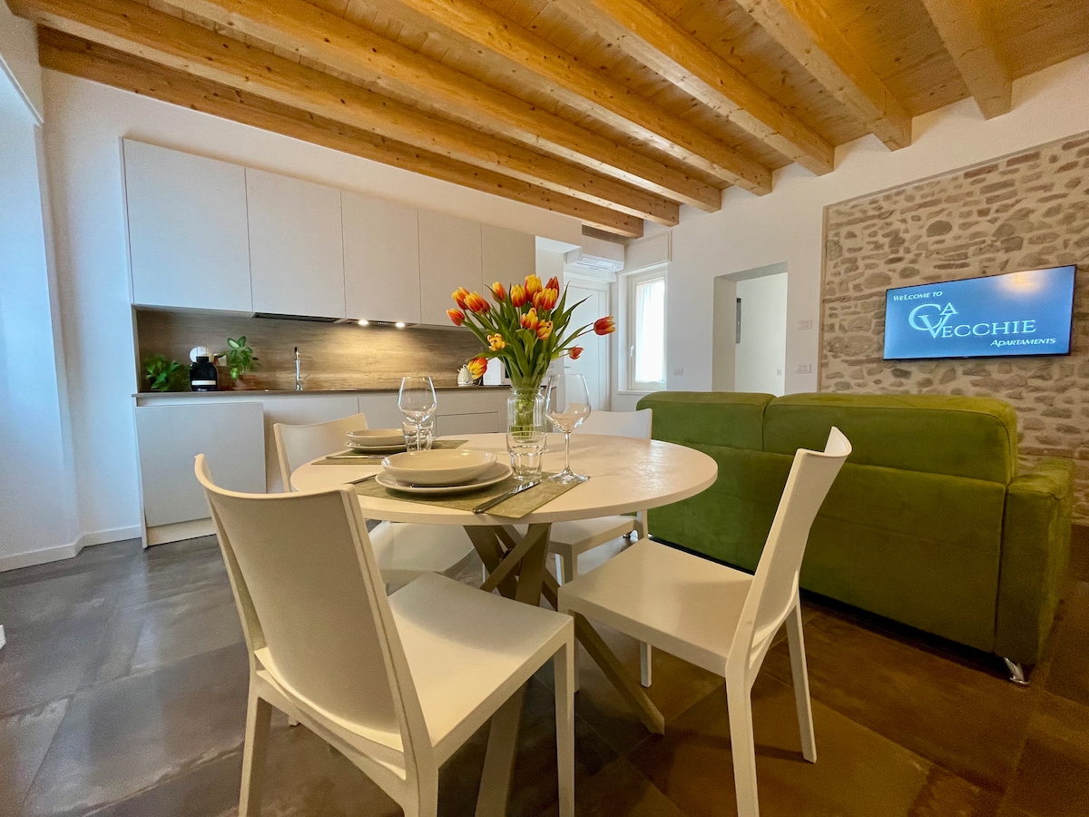 A modern dining area is set with a round table, surrounded by white chairs. A vibrant bouquet of tulips is displayed at the center. Natural light brightens the space, highlighting the wooden beams and the earthy tones of the stone wall in the background.