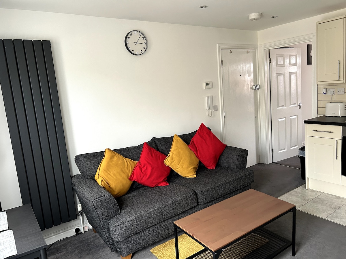 A modern living area features a gray sofa adorned with colorful red and yellow cushions. A small wooden coffee table sits in front, offering a simple space for relaxation. A wall clock is positioned above, and a doorway leads to an adjacent room.