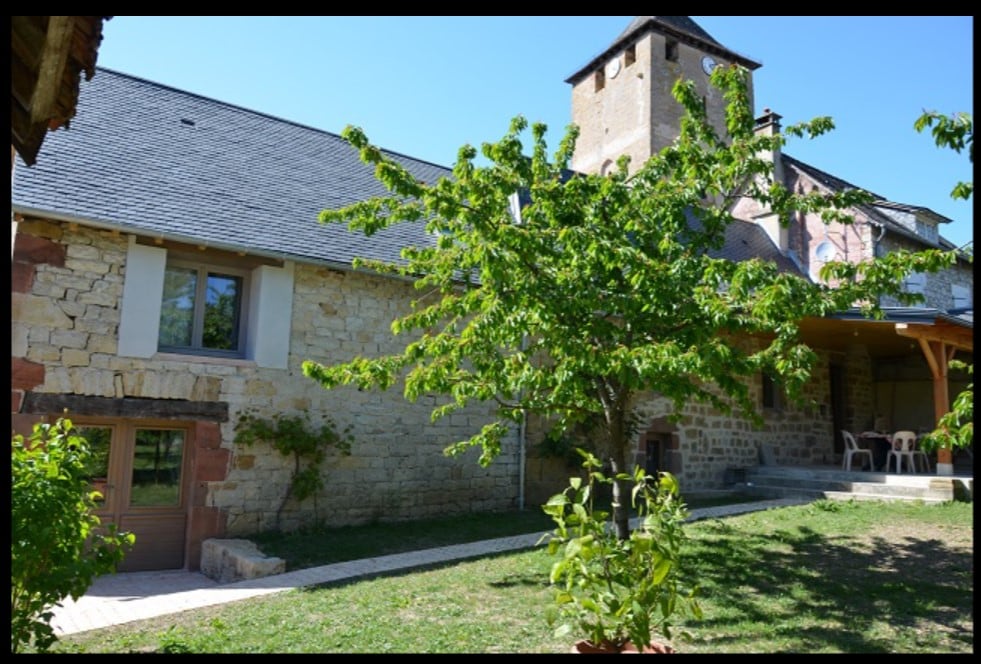 An ancient stone house stands within a serene garden, featuring a flat, fenced yard and a large terrace. A leafy tree provides shade, while neighboring structures exhibit rustic architecture. Multiple outdoor seating options are visible near the entrance, enhancing the inviting exterior ambiance.