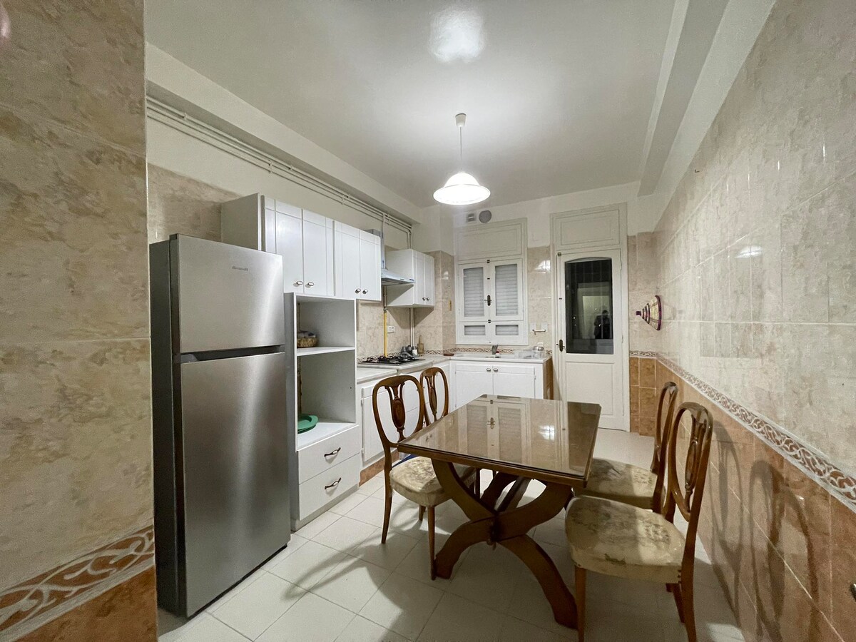 A modern kitchen adorned with light-colored tiles features a spacious dining table surrounded by four chairs. Stainless steel appliances, including a large refrigerator, complement the cabinetry, while natural light enters through a nearby window.