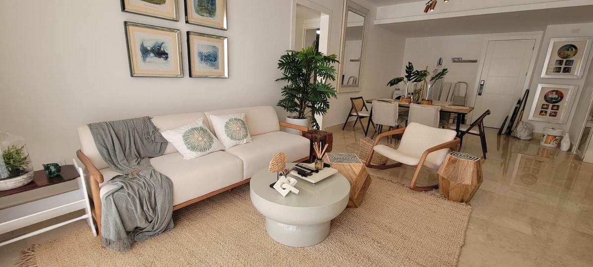 A spacious living area features a light-colored sofa adorned with decorative cushions. A round coffee table sits at its center, surrounded by natural-textured accents. In the background, a dining table is visible, complemented by modern chairs and potted plants that enhance the serene ambiance.