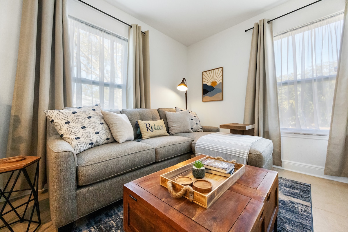 The living area features a comfortable gray sofa adorned with decorative pillows, alongside a wooden coffee table with serving trays. Two windows let in natural light, framed by soft curtains, while a piece of art on the wall adds a touch of personality.