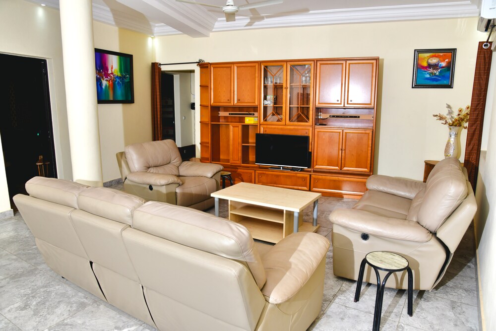 A spacious living area is furnished with comfortable leather seating arranged around a central coffee table. A wooden entertainment unit houses a television, with artwork displayed on the walls. Natural light is provided through nearby windows, enhancing the room's inviting atmosphere.