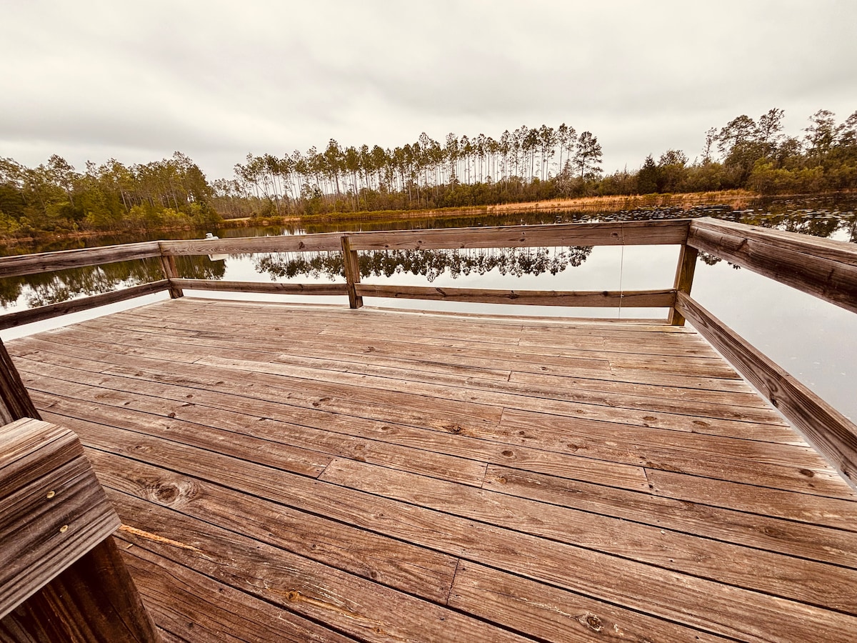A rustic wooden dock extends over a calm pond, surrounded by tall trees and lush greenery. The tranquil water reflects the cloudy sky and wooded landscape, creating a peaceful setting for relaxation or fishing.