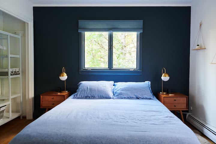 Თანამედროვე საძინებელი ფლოტის აქცენტის კედლით, კომფორტული თეთრეულითა და შუა საუკუნეების ტუმბოებით. Ფანჯარა ტყის ხედით. Ჩაშენებული კარადა. Მინიმალისტური დეკორი დასაკიდი თაროებით. Მყუდრო და ელეგანტური დიზაინი დასასვენებლად და მშვიდი ძილისთვის.