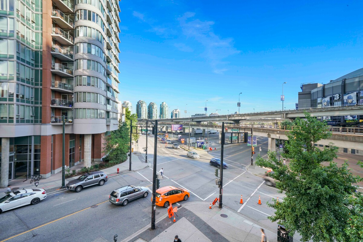 A view of downtown Vancouver showcases a bustling intersection with a mix of vehicles, including an orange car, traveling along the street. Towering buildings line the background, and a blue sky with sparse clouds frames the scene, emphasizing the urban environment.