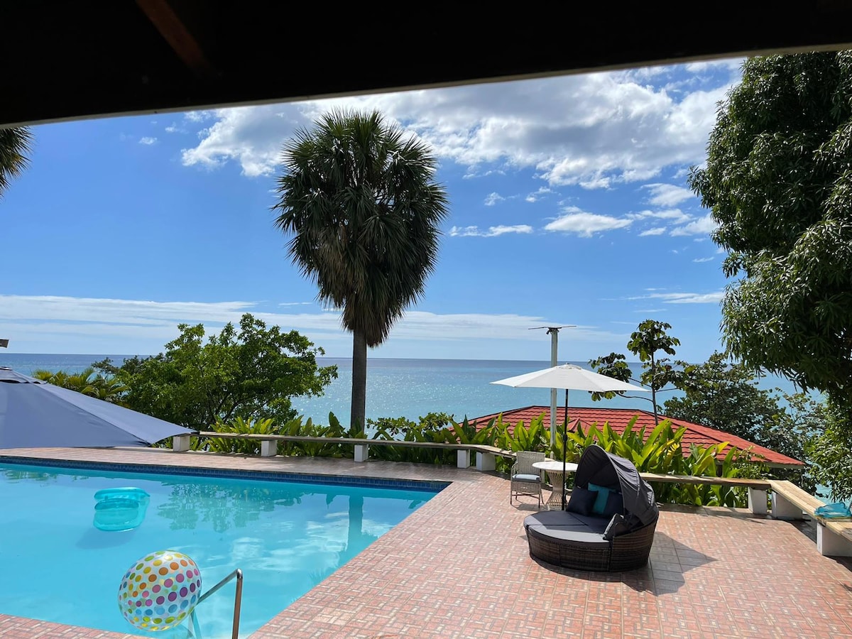 A spacious outdoor area features a sparkling pool, surrounded by lush greenery and trees. The view of the ocean is visible in the background, while a sun umbrella provides shade. A large lounge chair invites relaxation, creating a serene setting.