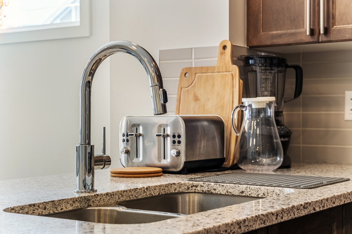 A modern kitchen countertop is showcased, featuring a sleek chrome faucet above a double sink. A stainless steel toaster sits beside a wooden cutting board and a blender, while a glass carafe and a dish drying mat are also visible.