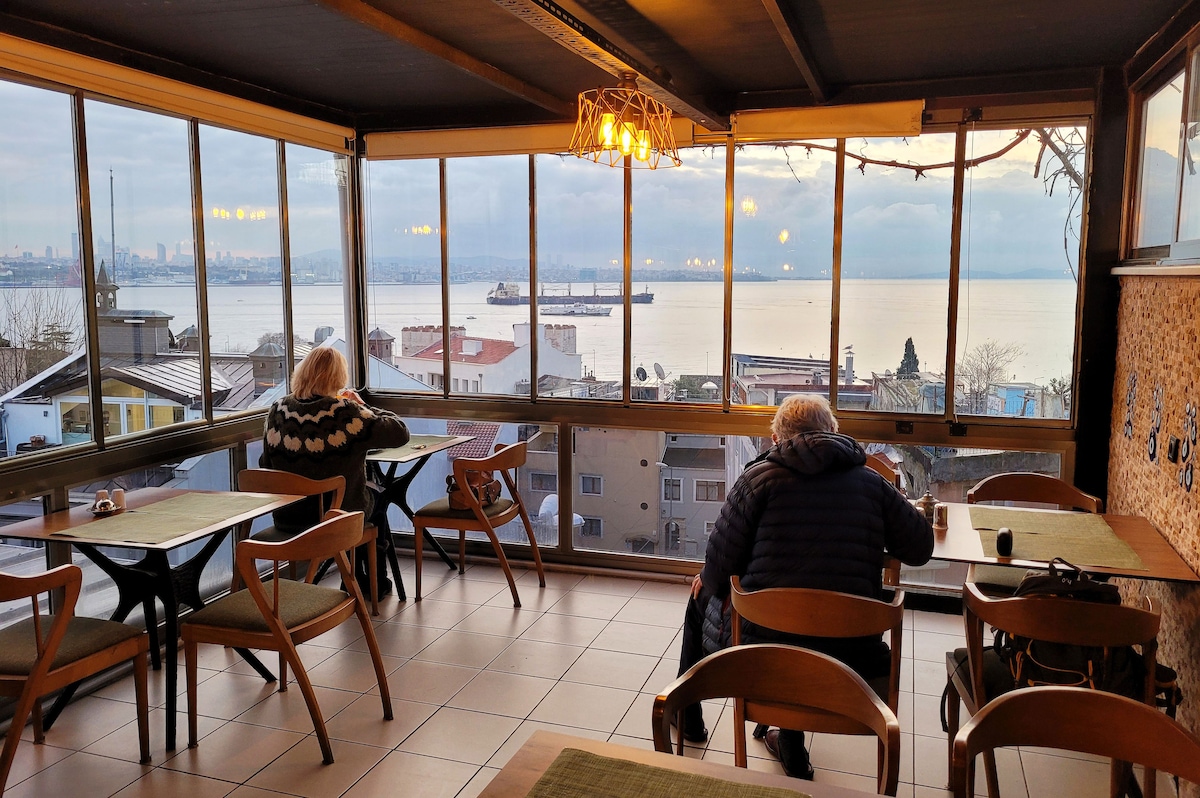 A sunlit dining area features large windows offering expansive views of the water and distant cityscape. Wooden tables with light green table settings are arranged to provide a comfortable atmosphere for guests enjoying their meals. Two individuals are seated quietly, engaging in conversation.
