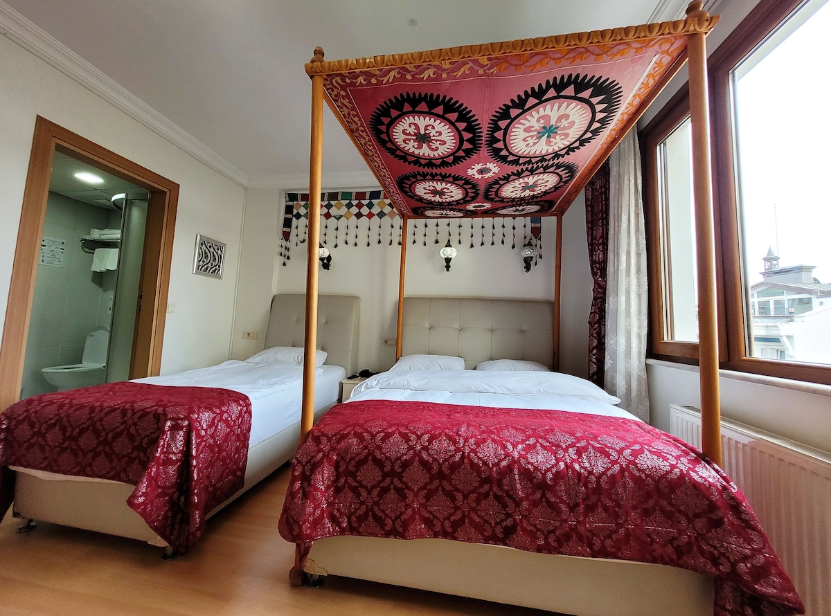 A guest room features two spacious beds adorned with rich red coverings. The canopy above includes a detailed patterned fabric. Natural light filters through large windows, illuminating the room's neutral tones and showcasing decorative wall elements.
