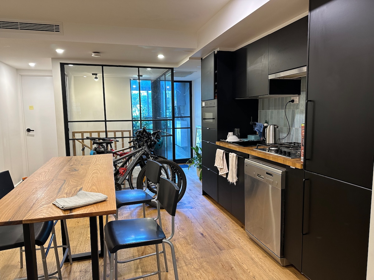 A modern kitchen area is featured with sleek black cabinetry and stainless steel appliances. A wooden dining table is complemented by black chairs, while bicycles are parked nearby, hinting at an active lifestyle. Natural light filters in through large glass doors.