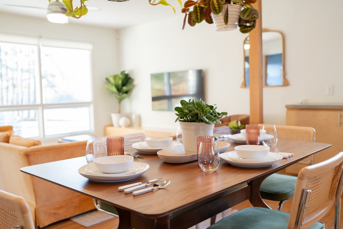 A dining table is set for four, featuring white plates, clear glasses, and woven napkins. A potted plant adds greenery to the center. In the background, a cozy seating area is visible, adorned with natural light filtering through large windows.