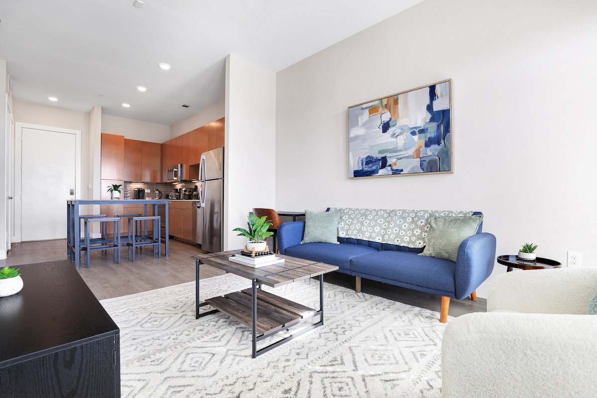 A spacious living area features modern furnishings, including a blue couch with decorative pillows and a coffee table. The kitchen with stainless steel appliances is visible in the background, along with a dining table and chairs. Neutral tones create a calming environment.