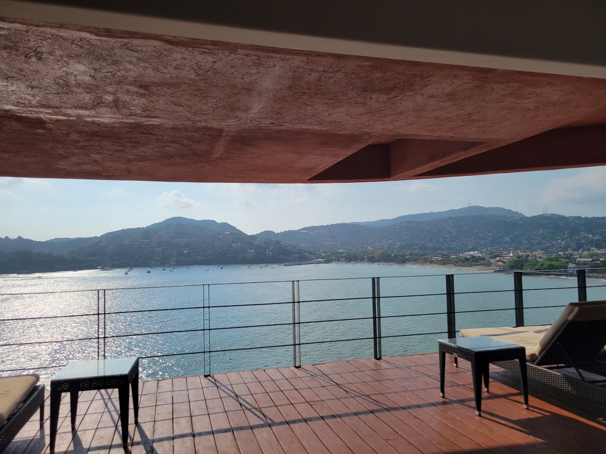 A spacious deck features sun loungers positioned for a panoramic view of the bay. Gentle waves reflect light from the sun, while distant hills frame the scenic backdrop, creating a tranquil setting.