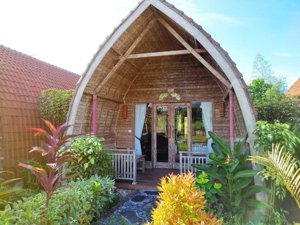 A charming cottage with a distinctive thatched roof is framed by lush greenery and colorful plants. Wooden doors and a cozy seating area are visible on the porch, inviting relaxation in the tranquil surroundings.