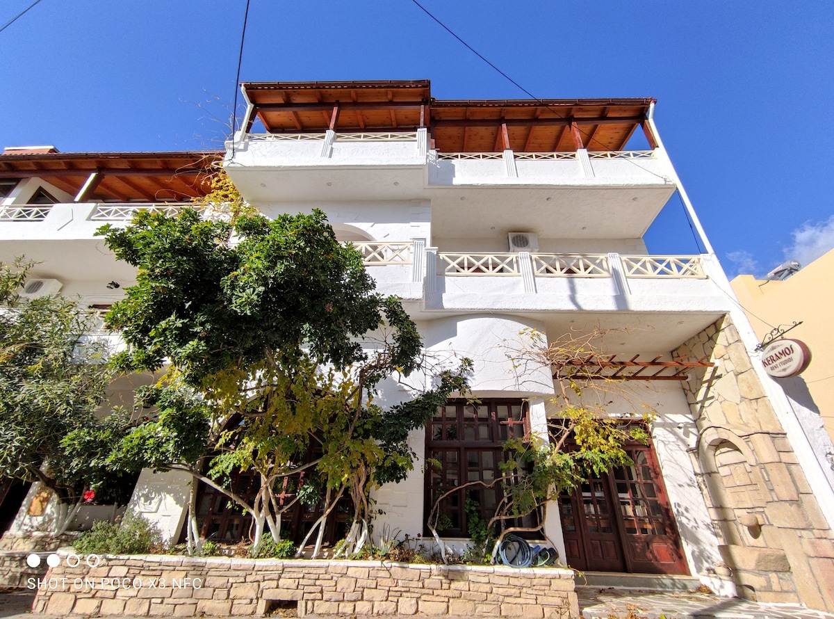 The charming guest house is displayed with a traditional design, featuring a white façade and wooden balconies. Lush greenery surrounds the entrance, creating a welcoming environment. Sunlight enhances the structure, while the blue sky provides a vibrant backdrop.