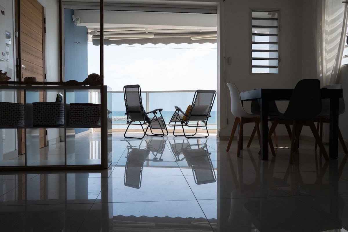 The room features a polished tile floor reflecting natural light. A glass door opens to a balcony with two loungers, offering a view of the sea. A dining table with white and black chairs is visible in the foreground, creating a sense of spaciousness.