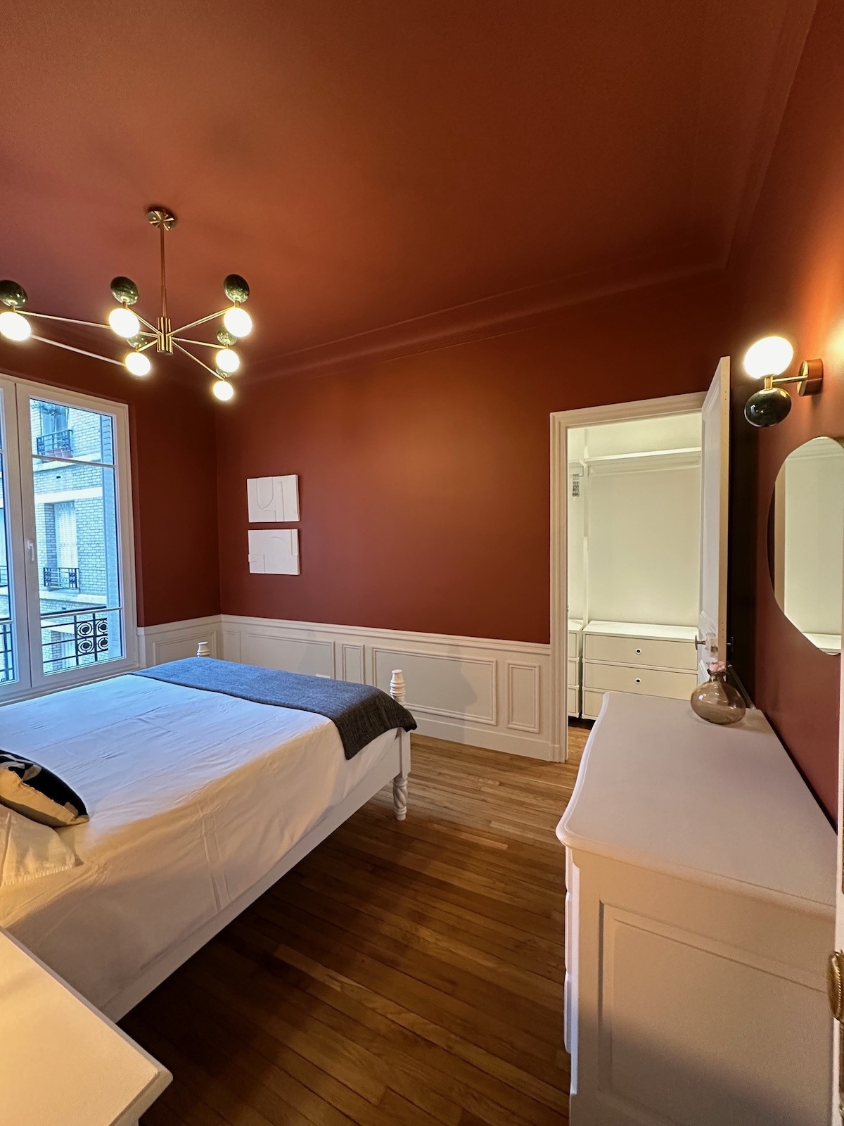 A well-lit bedroom features a spacious bed with a neutral-colored blanket. A stylish chandelier provides illumination from the ceiling. The deep red walls create a warm ambiance, and a large window allows natural light to fill the room. A dresser and a mirror are also visible.