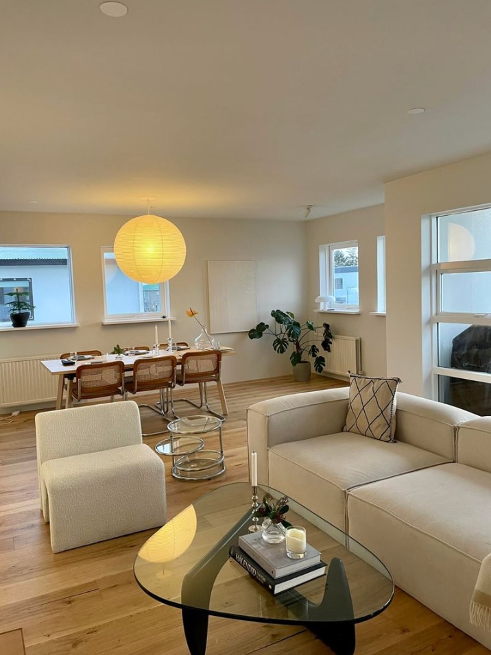 A modern living space is seen, featuring a light-colored sofa paired with a glass coffee table. A decorative lamp provides soft illumination. A dining area with wooden chairs is positioned in the background beside windows that allow for natural light.