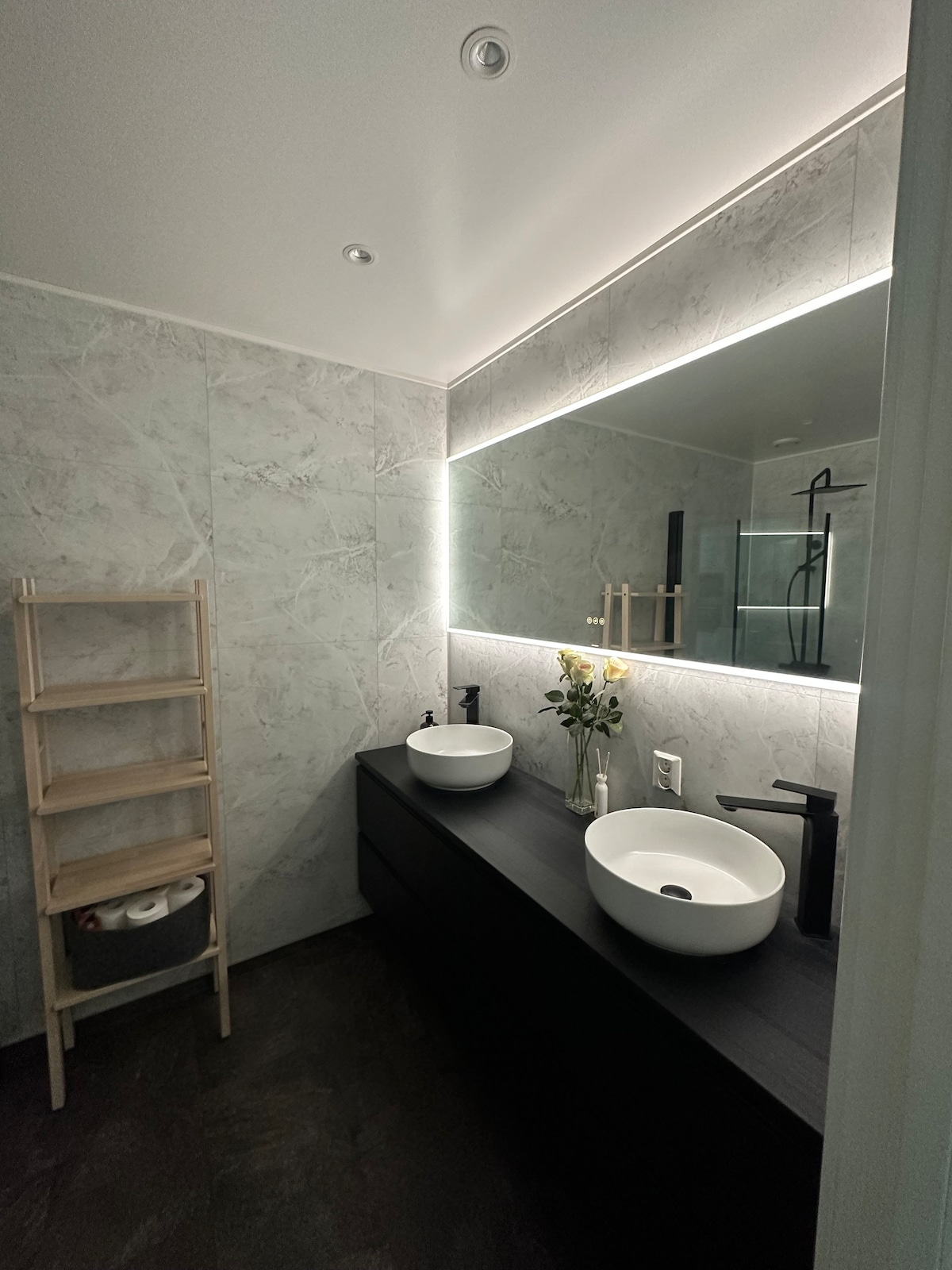 A modern bathroom features two round sinks on a sleek dark cabinet. Soft lighting illuminates the stone-patterned walls, enhancing the spacious feel. A minimalist wooden shelf stands beside the sinks, holding essential items. A glass shower enclosure is visible in the background.