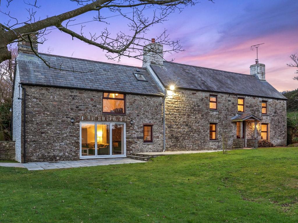 A charming farmhouse exterior is showcased, with a stone facade and multiple windows reflecting soft evening light. The property features a spacious patio area leading to a well-kept garden, surrounded by trees under a colorful twilight sky.