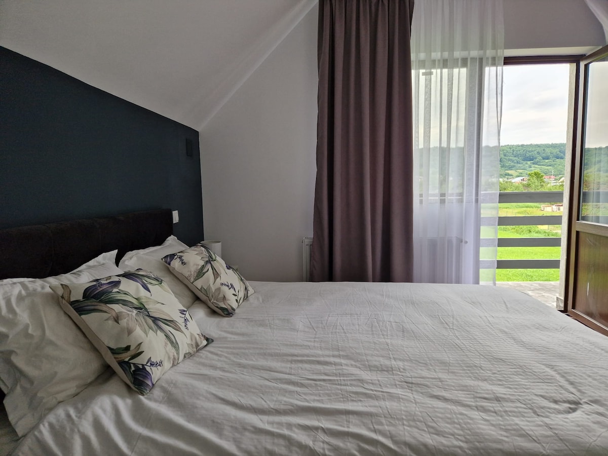 A serene bedroom is displayed, featuring a spacious bed adorned with light-colored bedding and decorative pillows. Natural light enters through an open window, illuminating the space and highlighting the green hillside view visible through the balcony railing.