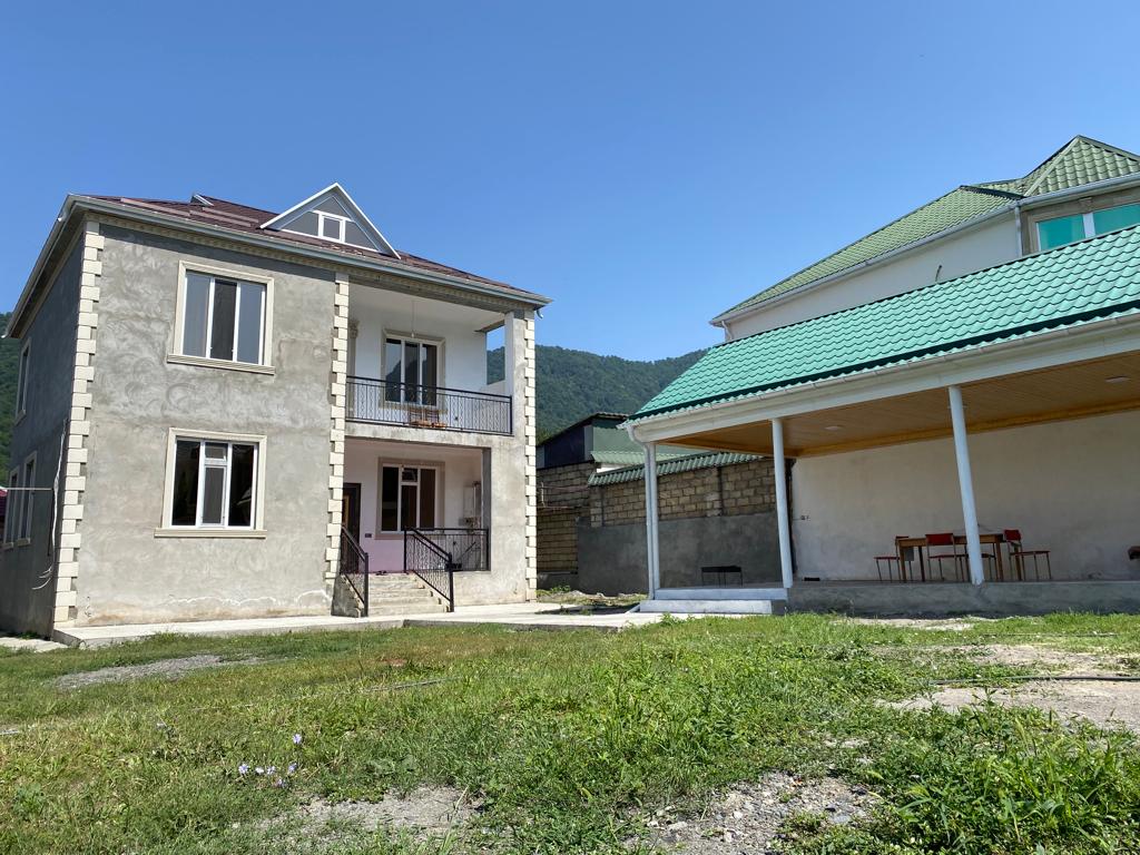 A two-story villa is depicted, featuring a blend of a stucco exterior and multiple windows that allow for ample natural light. A covered outdoor seating area is visible to one side, surrounded by grass. The backdrop includes green hills under a clear blue sky.
