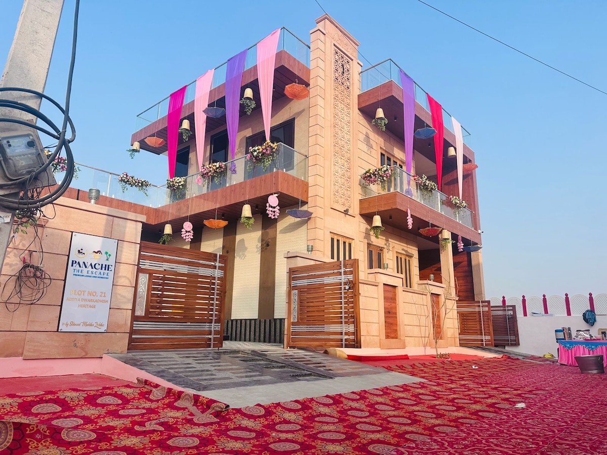 A stylish two-story building is adorned with colorful decorations, showcasing vibrant drapes and hanging flower arrangements. The welcoming entrance is framed by wooden gates, while the ground area is covered with a decorative rug. Clear skies complement the inviting atmosphere of the property.