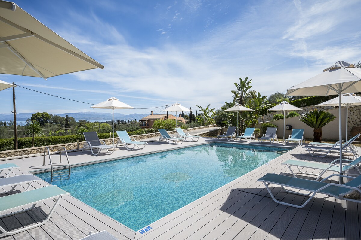 A well-maintained outdoor swimming pool is surrounded by several lounge chairs and umbrellas. Lush greenery and palm trees are visible in the background, while the sky displays a clear blue hue with soft clouds. The area provides a relaxing environment for leisure.
