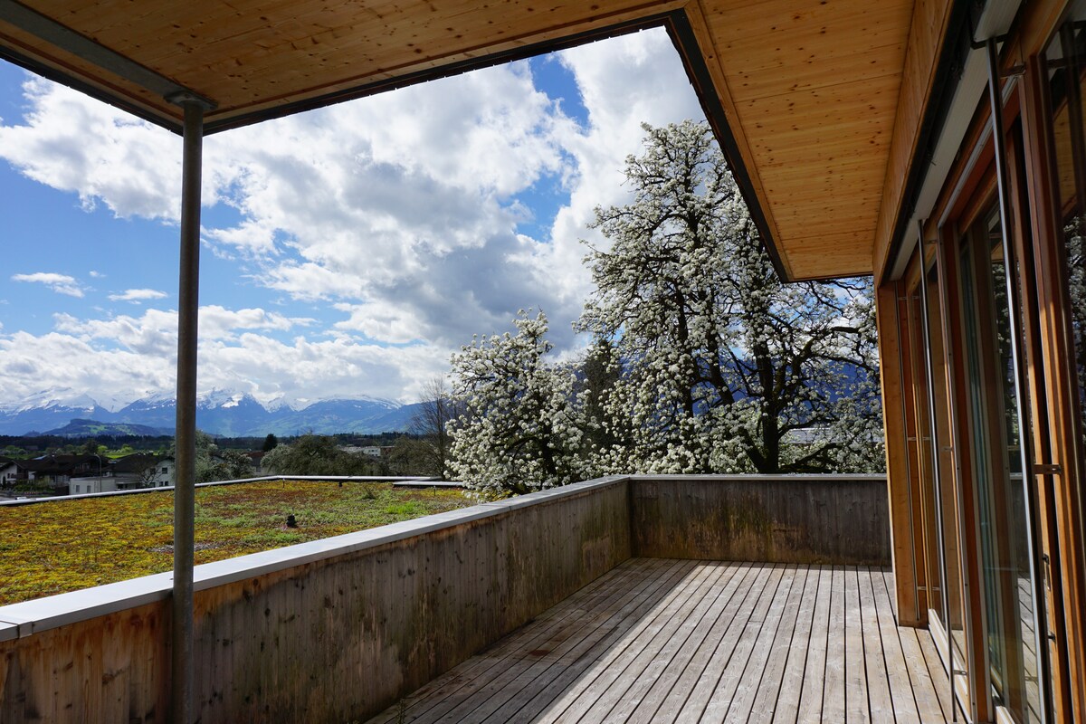A spacious terrace is highlighted by natural wood flooring and surrounded by greenery. Blossoming trees provide a vibrant contrast against the backdrop of distant mountains and a blue sky dotted with clouds. The open space invites fresh air and natural light.