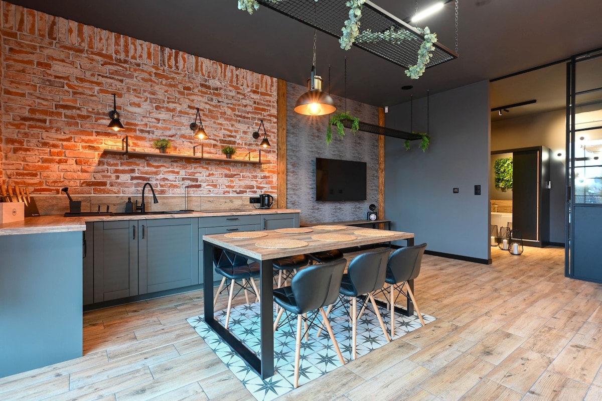 An open-plan kitchen and dining area is presented, featuring a wooden dining table accompanied by four black chairs. Exposed brick walls are complemented by modern lighting fixtures. A flat-screen television is mounted on the wall, and a kitchenette is visible to one side.