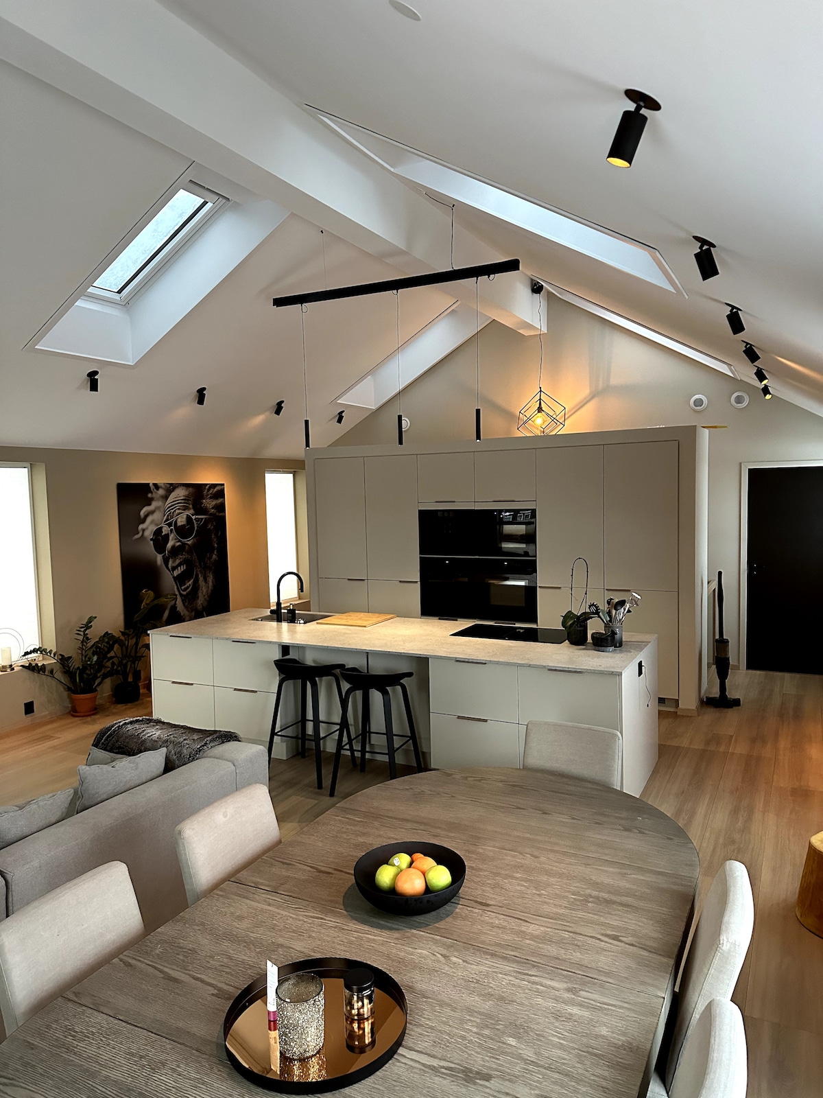 A modern kitchen is showcased with sleek cabinetry and a large central island. Skylights allow natural light to fill the space, highlighting a dining table set with a circular tray of fruits. Black bar stools are positioned at the island, inviting casual meals.