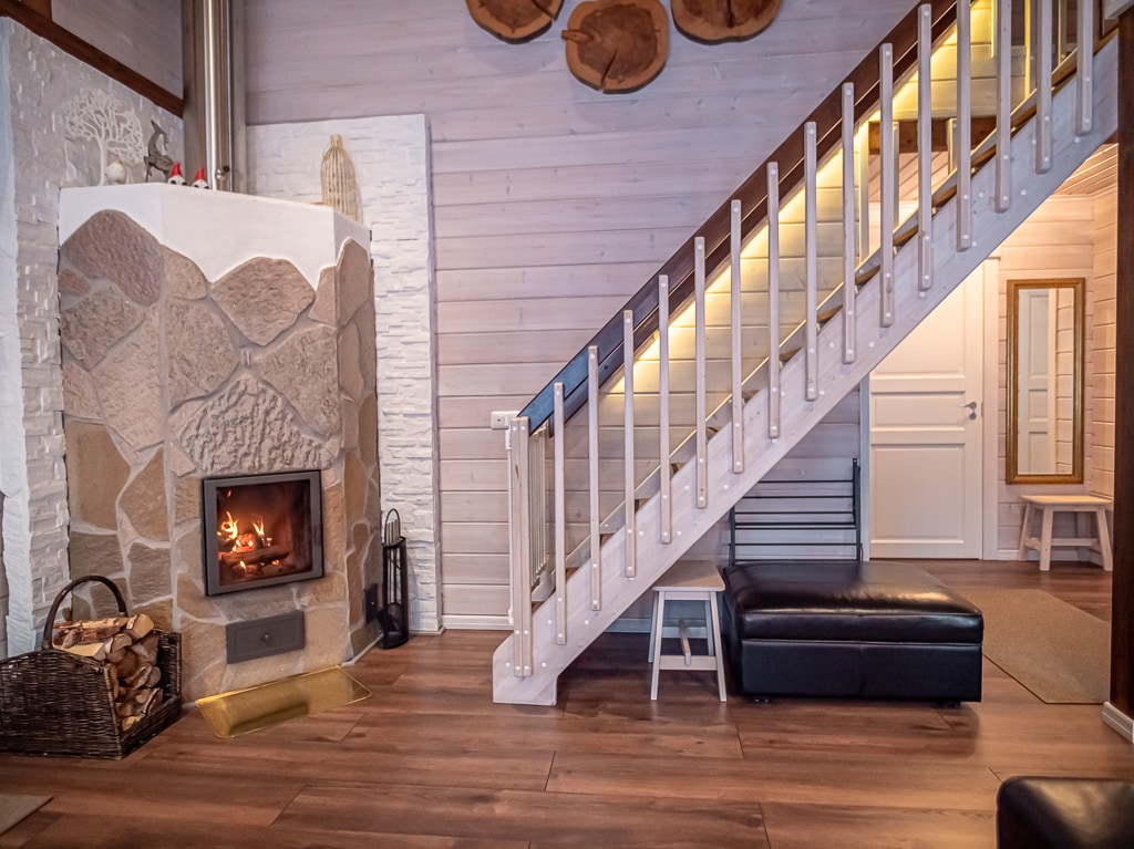 The living area features a stone fireplace with a warm fire, providing a cozy focal point. Light wood walls create an inviting atmosphere. A staircase leads to an upper level, while a black leather sofa adds comfort to the space. The area is brightened by soft lighting.