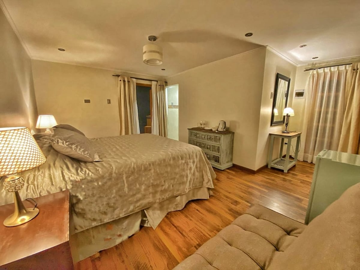 A spacious bedroom is presented, featuring a large bed with a soft, light-colored coverlet. A bedside lamp illuminates a wooden nightstand. A comfortable seating area includes a neutral-toned sofa. Light-colored curtains frame a window, enhancing the serene atmosphere of the space.