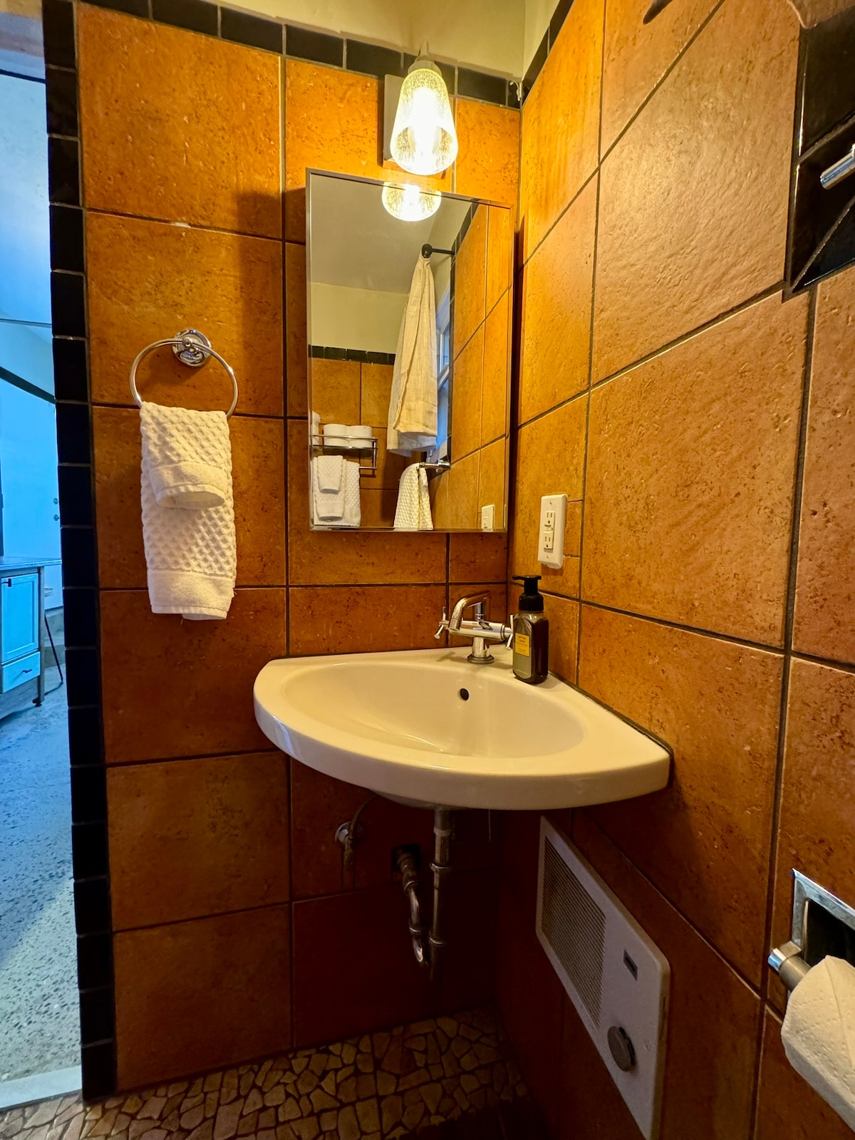 A compact bathroom features warm, earthy-toned walls and a modern sink with an attached mirror. White towels hang neatly on a towel rack. A light fixture above provides illumination, creating a functional and visually cohesive space.