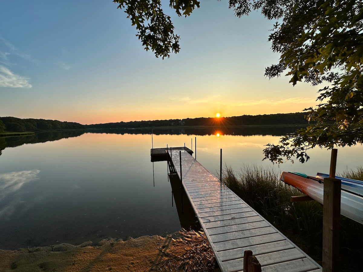 A serene view of a dock extending over calm waters during sunset, with reflections of trees and the sky visible on the surface. The landscape is framed by lush greenery on both sides, creating a peaceful and inviting atmosphere.