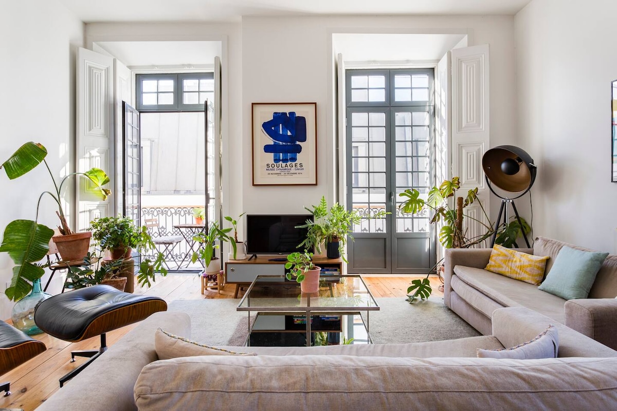 A sunlit living room features large open windows that invite natural light. Indoor plants adorn the space, complementing neutral-colored sofas and a glass coffee table. A modern art piece and a stylish lamp enhance the contemporary atmosphere, promoting a relaxed and inviting setting.