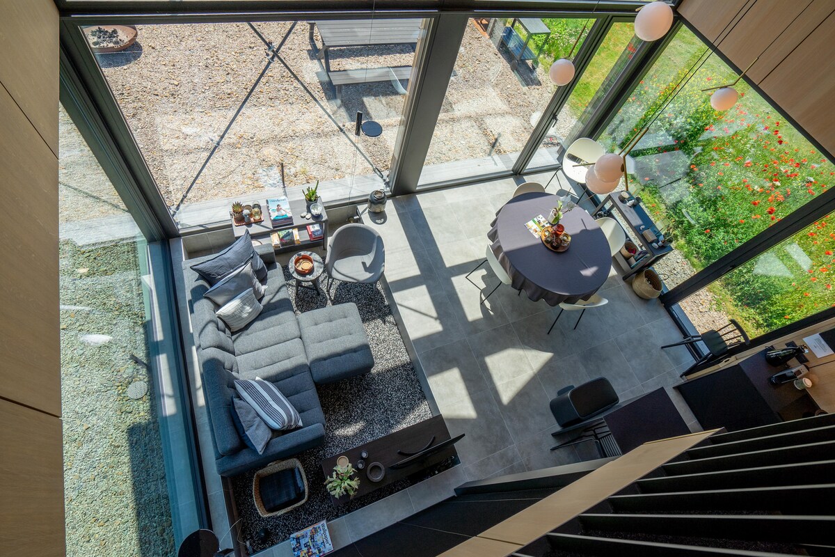 An aerial view reveals an open living space featuring a modern sofa and a round dining table. Large glass walls invite natural light, while greenery is visible outside. Decorative items can be seen on coffee tables, providing a glimpse of a harmonious indoor-outdoor atmosphere.