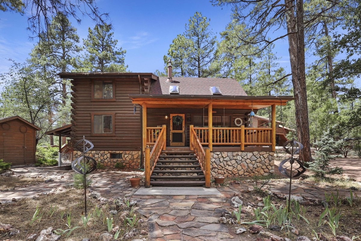 The cabin's exterior is framed by towering pine trees, featuring a welcoming front porch with a wooden railing. Natural stone accents complement the log-style architecture. Steps lead up to the entrance, inviting guests to explore the surroundings of the serene forest.