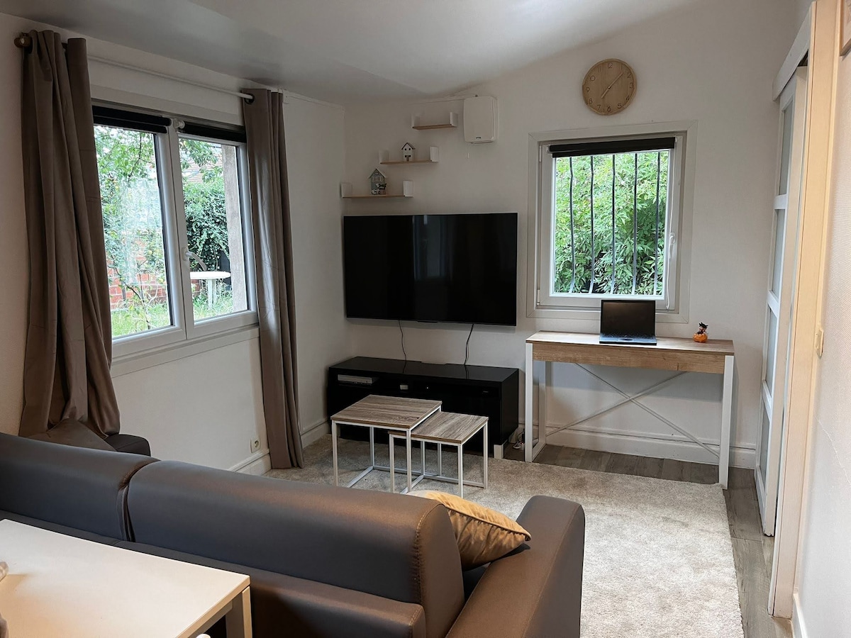 A comfortable living area features a large television mounted on the wall, with a minimalist black media console beneath. A small desk is positioned nearby, accompanied by a chair. Light streams through two windows, framed with neutral curtains, revealing green foliage outside.