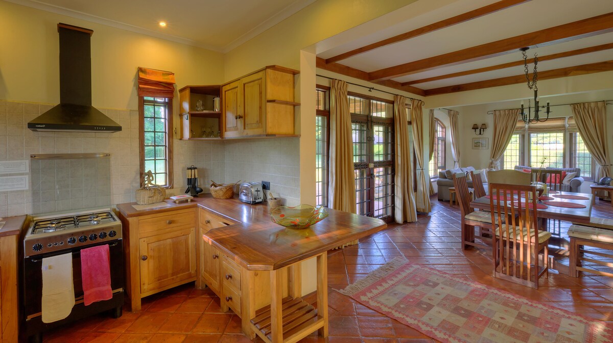 An open kitchen area features wooden cabinetry and a gas stove. A central island provides additional preparation space. Beyond, a dining area with a round table and wooden chairs is visible. Large windows allow natural light, highlighting the tiled floor and rustic decor.