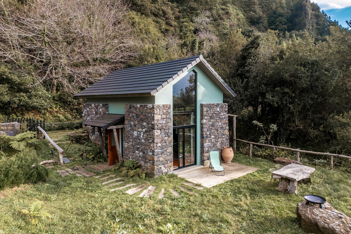A charming house is nestled among lush greenery, featuring natural stone walls and a sloped roof. Large glass doors create a seamless connection to the outdoors. A wooden pathway leads to a simple outdoor seating area with a stone table, surrounded by the tranquil landscape.
