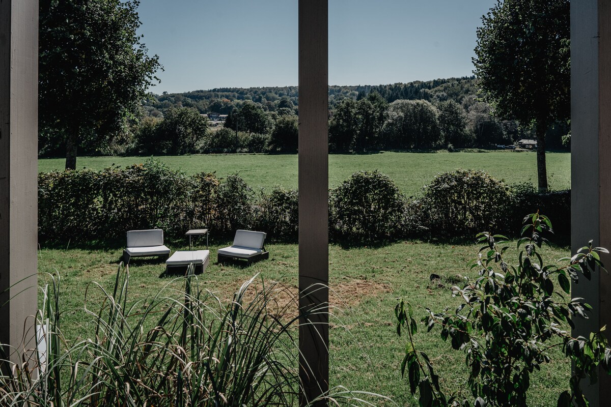 A view from the Tinyhouse shows a large green lawn surrounded by trees and shrubs. Two lounge chairs and a small table are positioned on the grass, inviting relaxation. The landscape extends into the distance, offering a peaceful rural setting.
