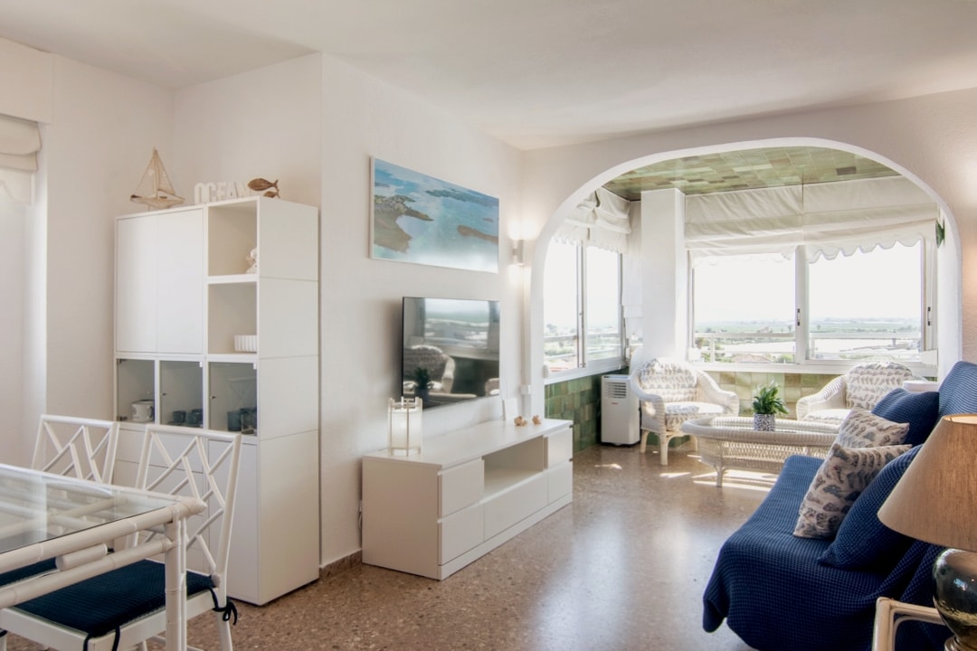 A bright living space features a comfortable seating area with blue cushions and a coffee table. Large windows offer expansive views of the surrounding landscape. A dining table with white chairs and a stylish shelf are also present, enhancing the open and airy atmosphere.