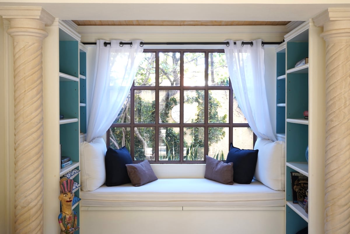 A cozy nook is created by a window seat adorned with white cushions and dark throw pillows. Flanking the seat are custom shelves painted in a light blue, showcasing books and decorative items. Natural light filters through sheer curtains, illuminating the inviting space.