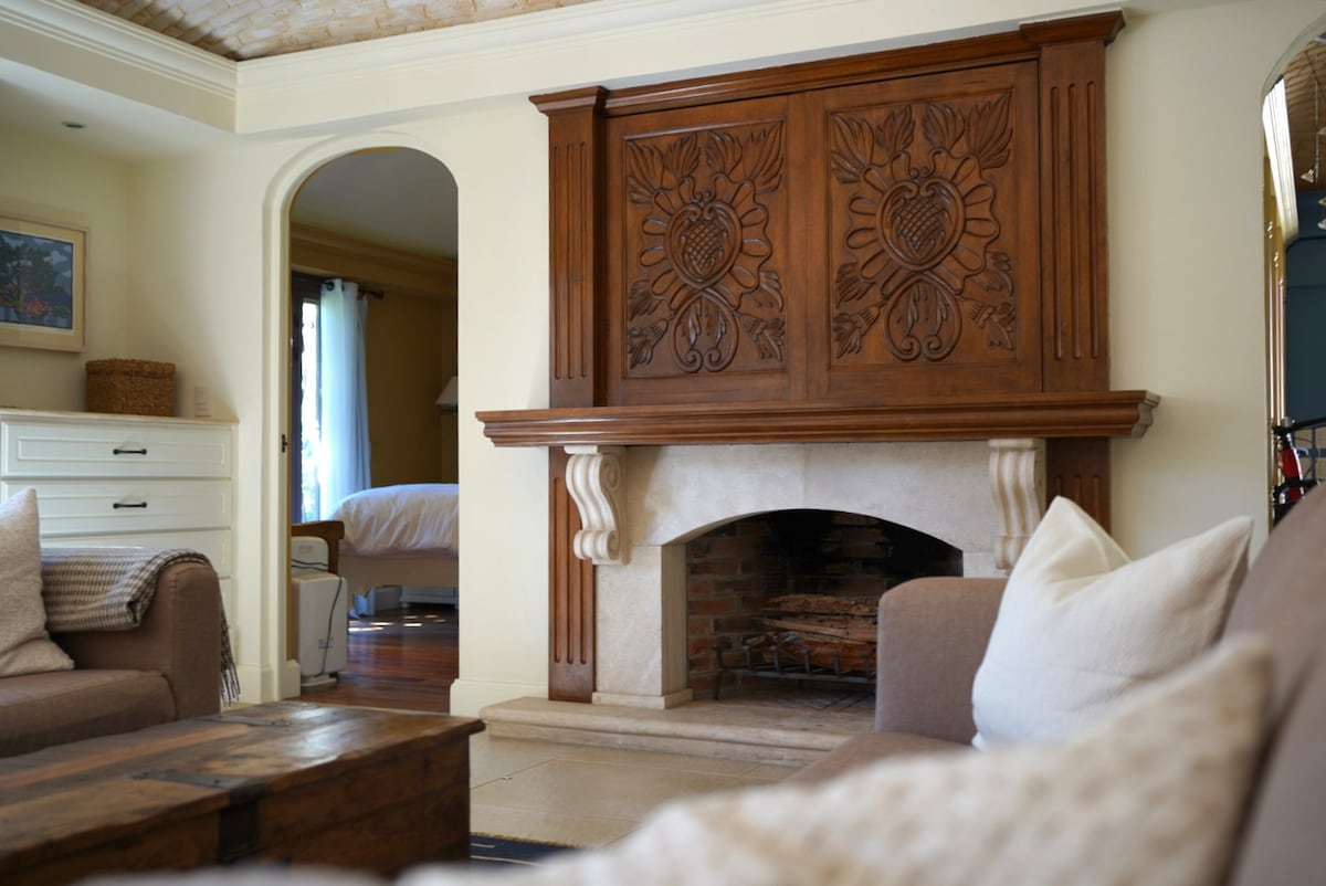 A warm living area features a carved wooden fireplace mantel, detailed with floral motifs. Beige couches with light-colored throw pillows create a cozy seating arrangement. An arched doorway leads to another room, while natural light illuminates the space.
