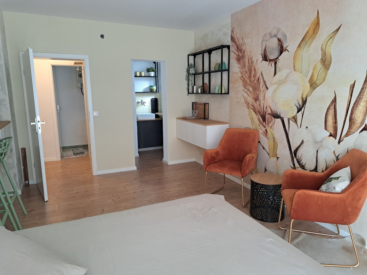 A living space features a stylish mural with cotton plant designs on a pastel wall. Two orange armchairs are positioned beside a small black side table, while a white console is mounted on the wall. A doorway leads to a kitchenette, enhancing accessibility and flow.