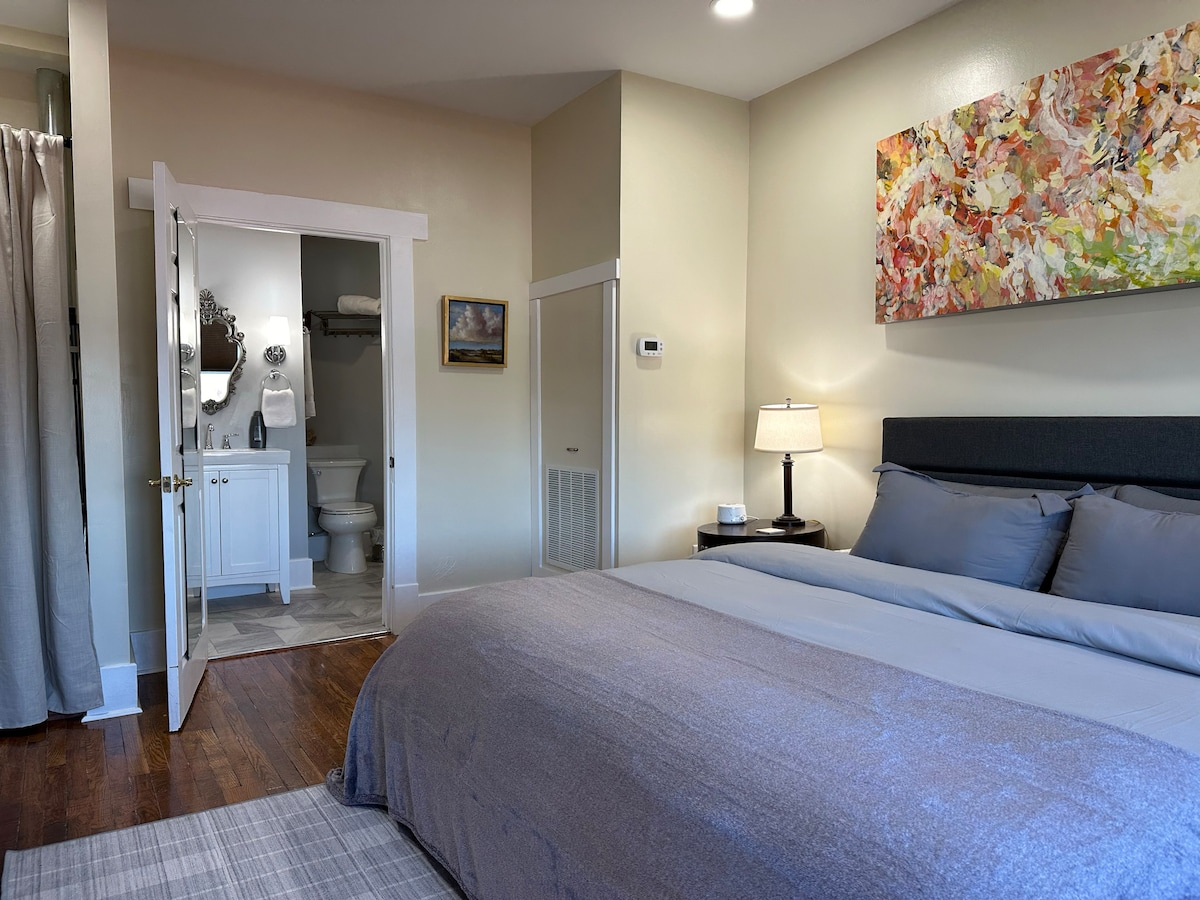 A bedroom features a spacious bed with soft grey bedding, complemented by a stylish painting above. A doorway leads to an attached bathroom, which is partially visible. Natural light brightens the room, enhancing the light-colored walls and wooden flooring.