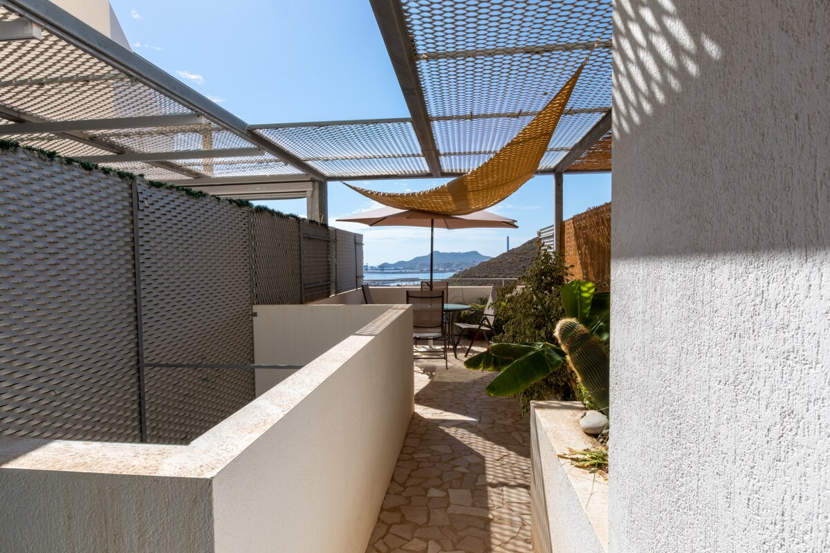 An outdoor path is framed by sleek stone walls, leading to a terrace shaded by a large, stylish awning. Comfortable seating arrangements are visible, surrounded by greenery, providing a serene setting with views of the distant sea.